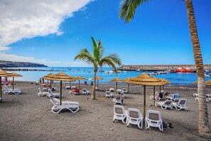 Una spiaggia con lettini e ombrelloni, un cielo azzurro limpido e barche sull'acqua.
