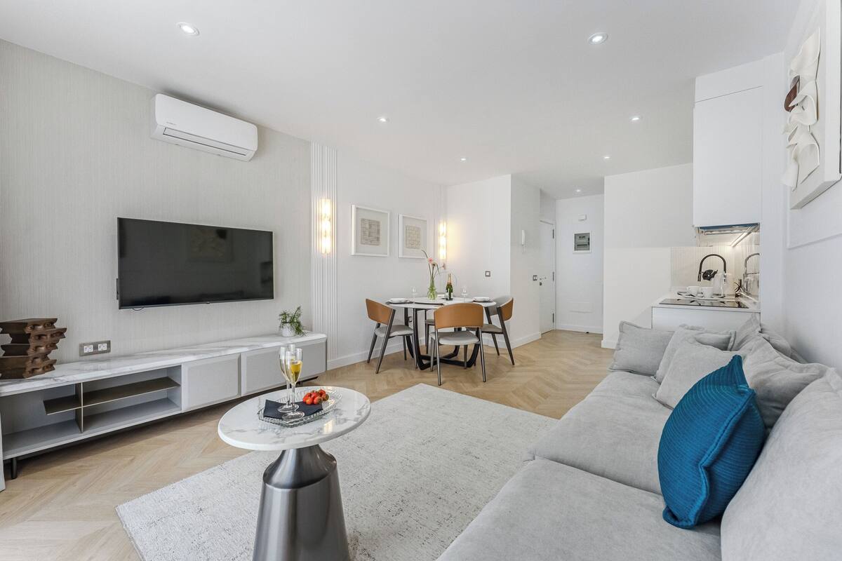 Ein modernes Wohnzimmer mit einem Flachbildfernseher, einem Essbereich mit Tisch und Stühlen sowie einem grauen Sofa mit blauem Kissen.