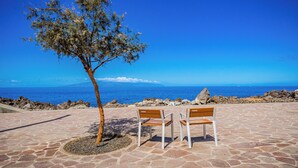 Ein mit Steinplatten belegter Bereich mit zwei hölzernen Bänken und einem Baum, mit Blick auf das klare, blaue Meer.