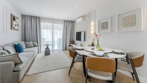 Ein modernes Wohnzimmer mit einer Couch, einem Esstisch und Stühlen. An der Wand ist ein Fernseher und ein Fenster mit Vorhängen.