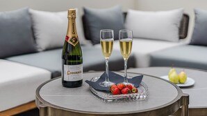 Une bouteille de vin Cuvée Original Brut, deux verres à vin, un plateau de fraises et deux poires, le tout posé sur une table ronde dans un salon moderne avec des canapés et des coussins gris.