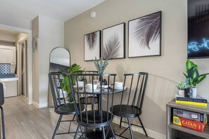 Un coin repas avec une table ronde, des chaises noires et une plante en pot. Des œuvres d’art encadrées ornent le mur, et une petite étagère contient des livres et une plante.