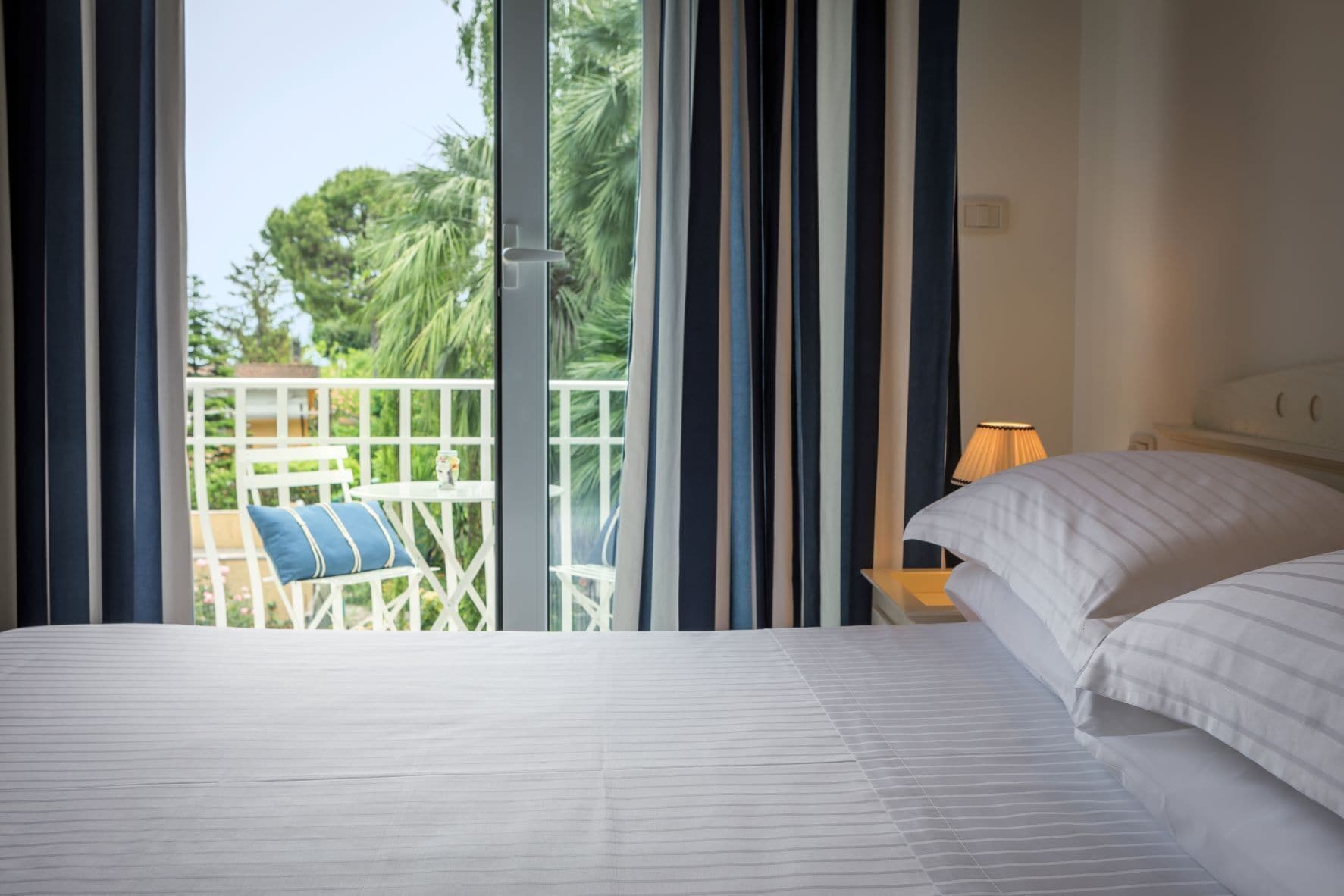 Une chambre avec un grand lit, une table de chevet, une lampe et une vue sur un balcon équipé d’une table et de chaises.