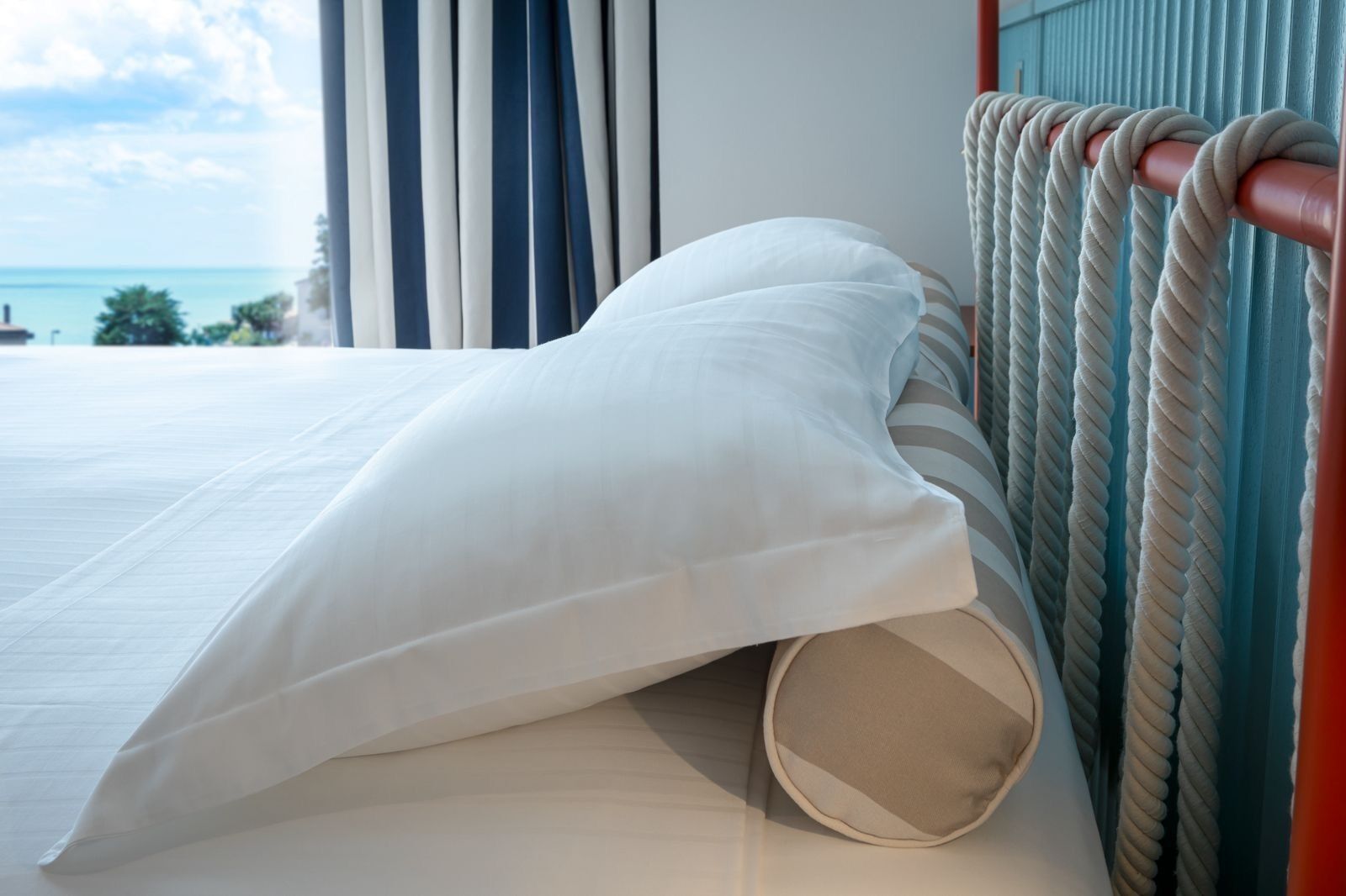 A bed with white linens, a view of the sea through curtains, and a rope-patterned headboard.