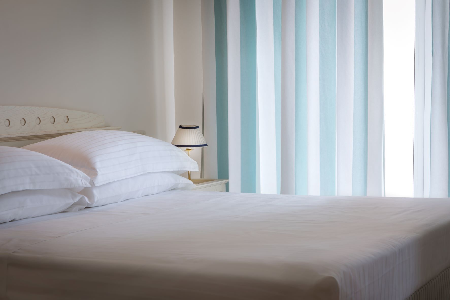Una cama bien hecha con sábanas blancas, una lámpara de noche y vistas a persianas verticales.