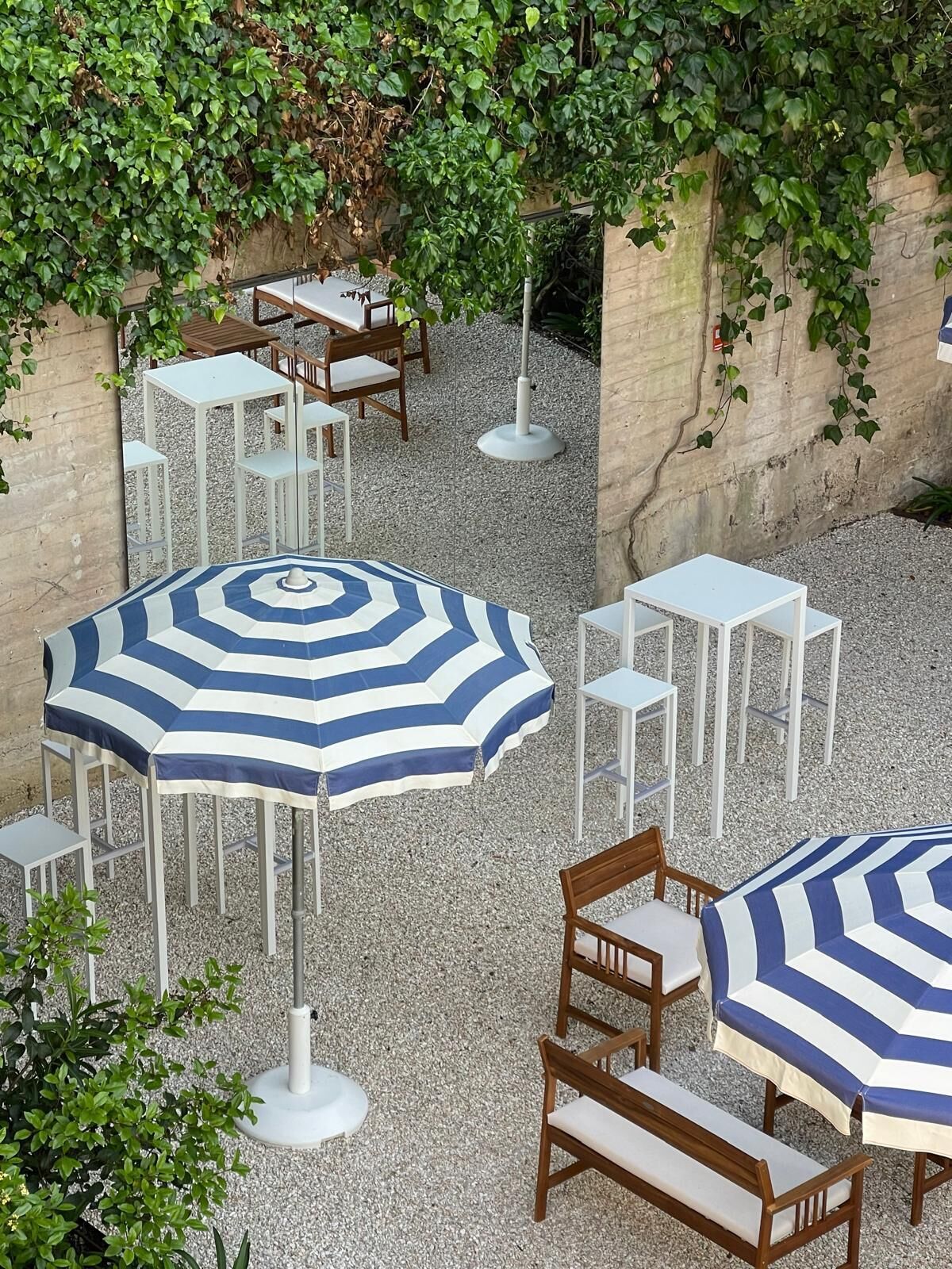 An outdoor seating area with tables and chairs, surrounded by greenery and a stone wall.