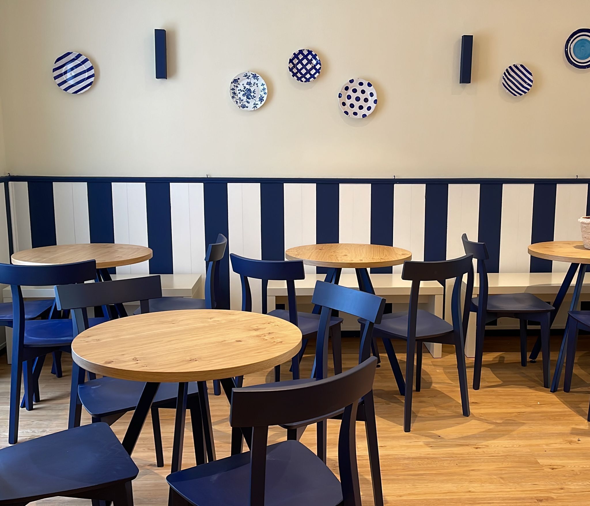A restaurant interior with round wooden tables and blue chairs, featuring a striped wall and decorative plates above the tables.