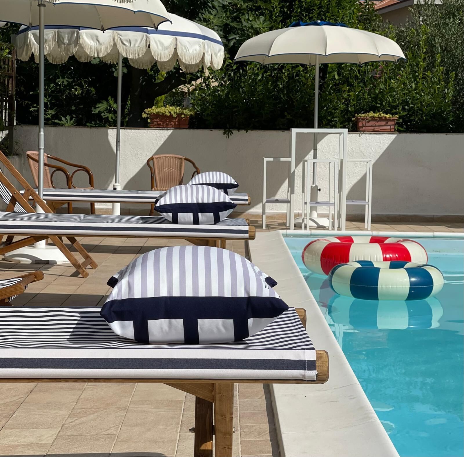A poolside area with lounge chairs and striped pillows, a pool with floating rings, and umbrellas.