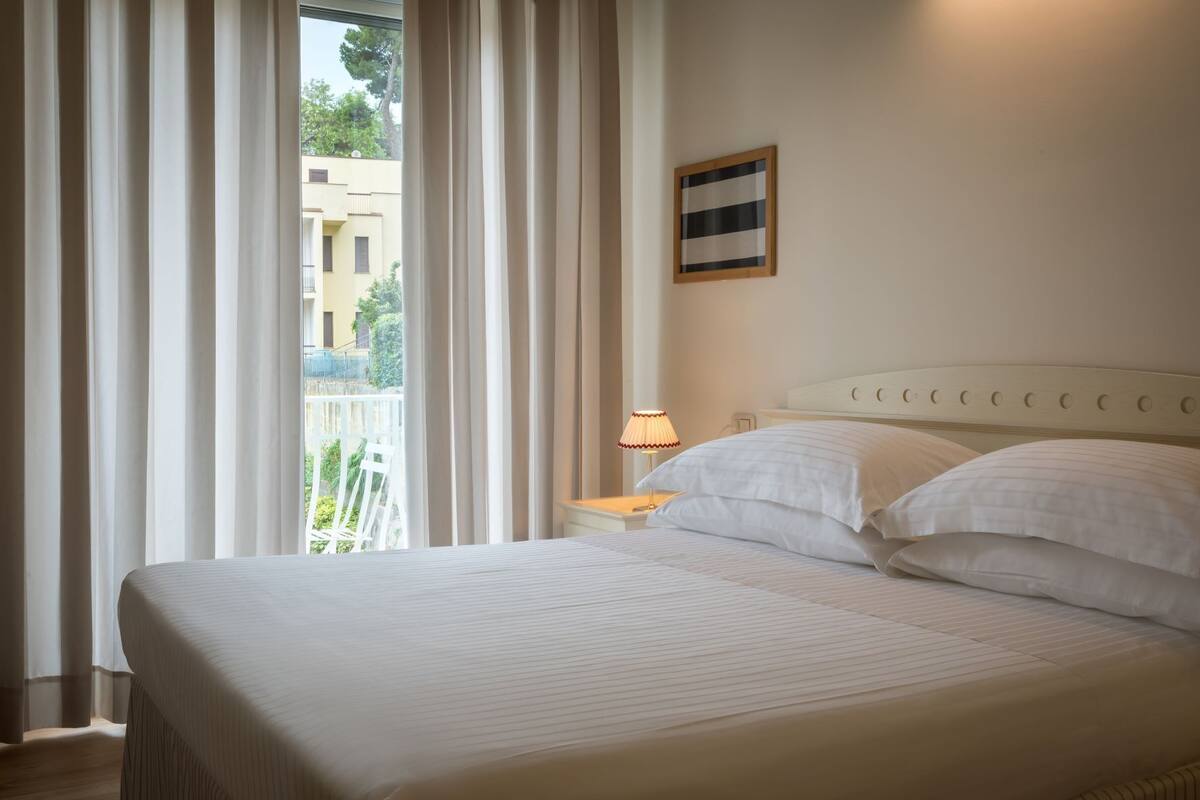 A hotel room with a large bed, a nightstand with a lamp, and a view of buildings through sheer curtains.