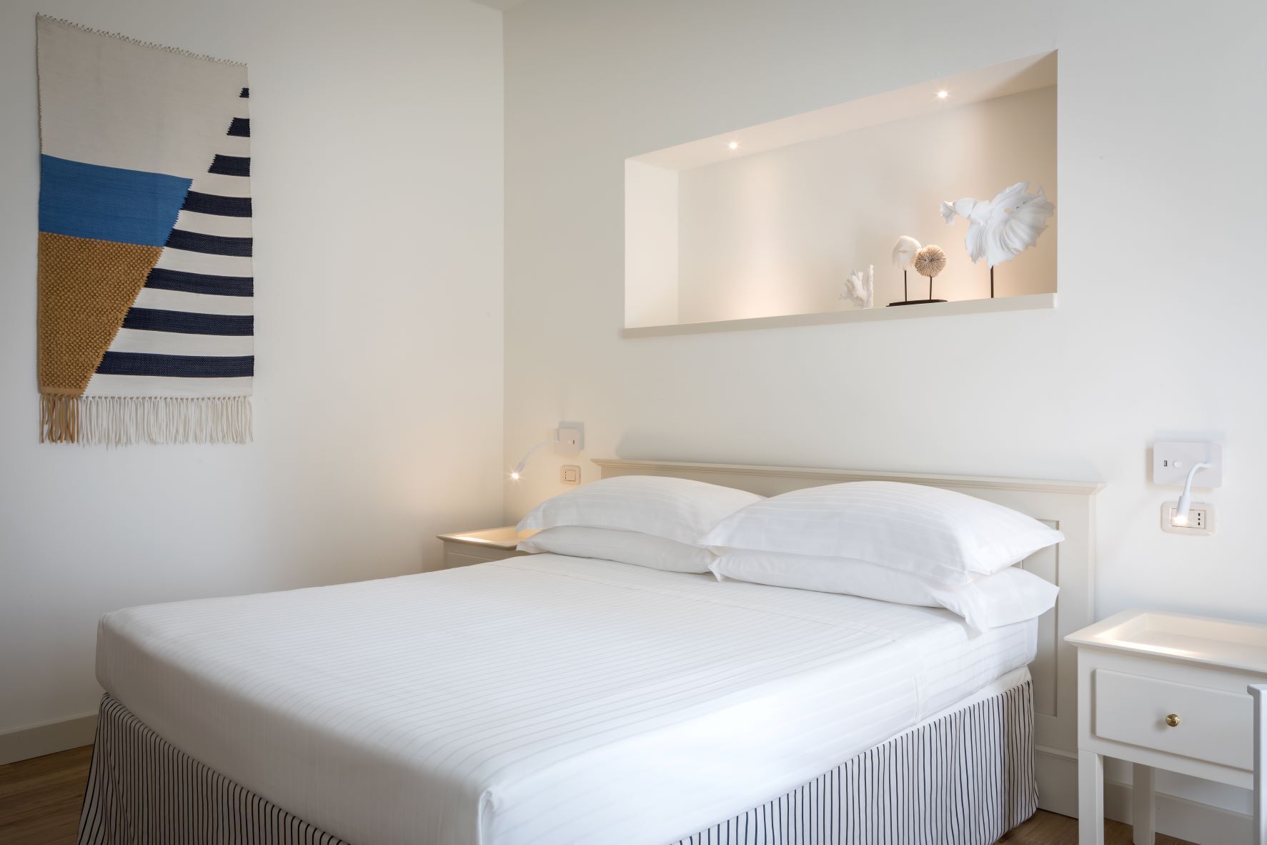 A neatly made bed with white linens, a bedside table with a lamp, and a wall-mounted shelf with decorative items.