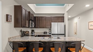 A kitchen with dark wood cabinets, a granite countertop, a stainless steel refrigerator, and a built-in island with bar stools. - Beach, Swim, Chill, Repeat – Alerio E202 (Miramar Beach)