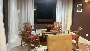 Ein gemütlicher Patio-Bereich mit Holzsesseln und einem Tisch, weißen Vorhängen und Blick auf die beleuchtete Stadt.