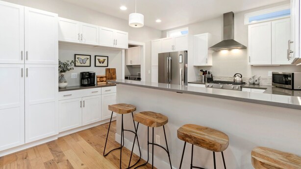 A modern kitchen with white cabinets, a stainless steel refrigerator, a gas stove, and wooden stools. - Ram’s Roost | Rooftop Hot Tub, Patio & Family Fun (Fort Collins)