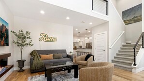 A modern living room with a sofa, armchair, and coffee table. There is a fireplace, a plant, and a wall-mounted sign. A staircase leads to an upper level. - Ram’s Roost | Rooftop Hot Tub, Patio & Family Fun (Fort Collins)