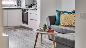 A modern living room with a grey sofa, a coffee table with books and a plant, and a kitchen in the background.