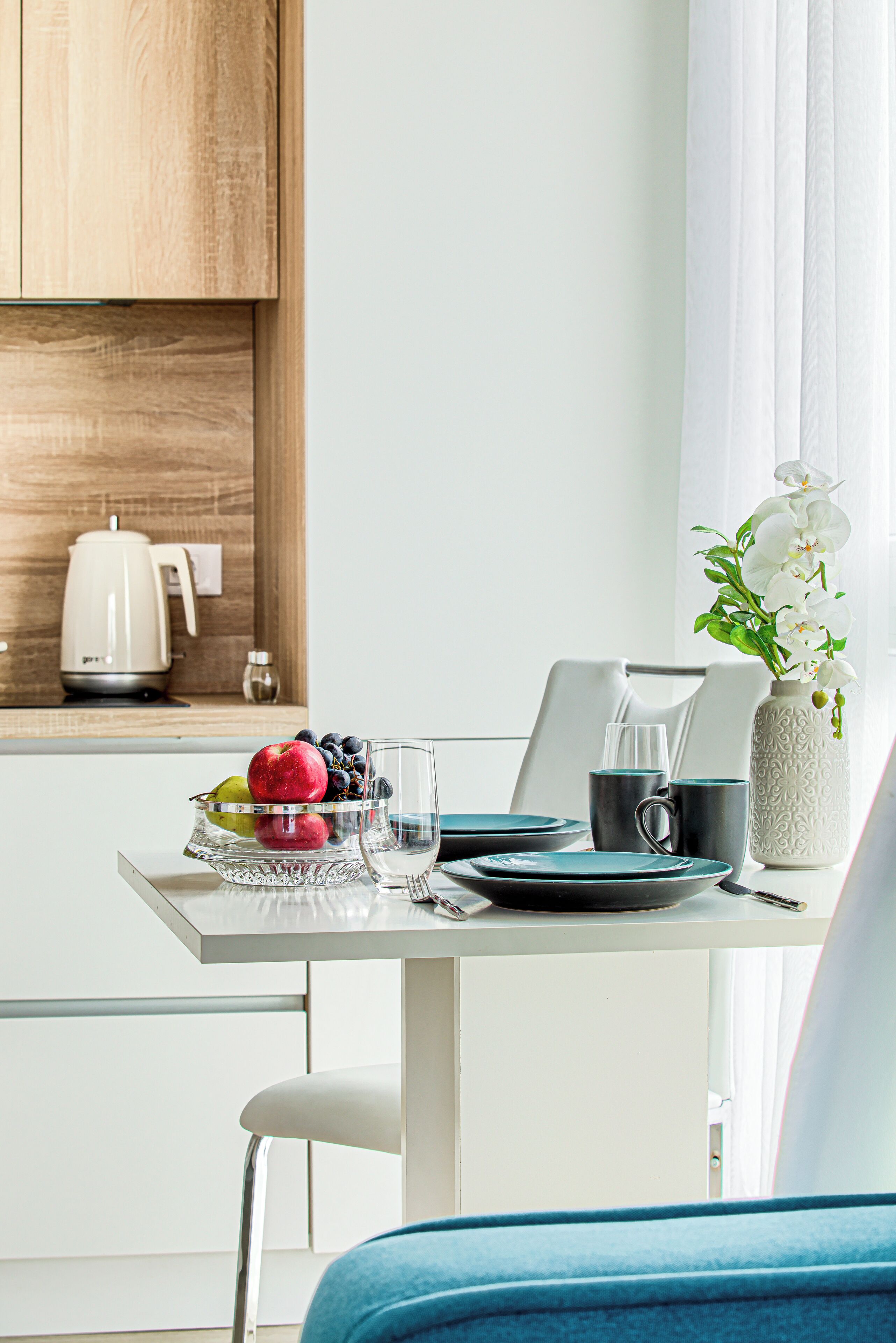 A modern kitchen with a white countertop, a kettle, a bowl of fruit, and a vase with flowers.