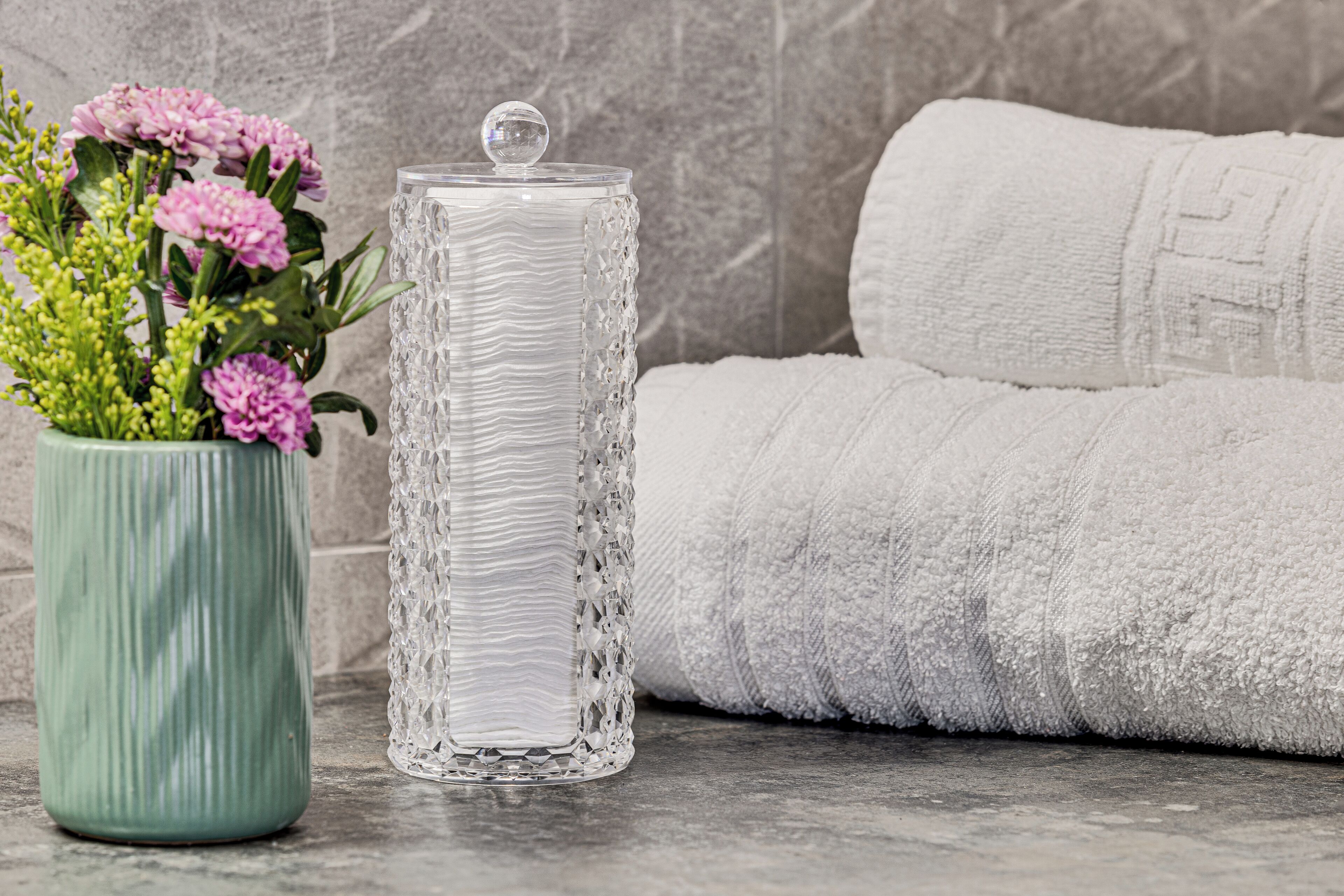 Un contenitore di vetro trasparente con coperchio, un vaso verde con fiori rosa e un asciugamano bianco.