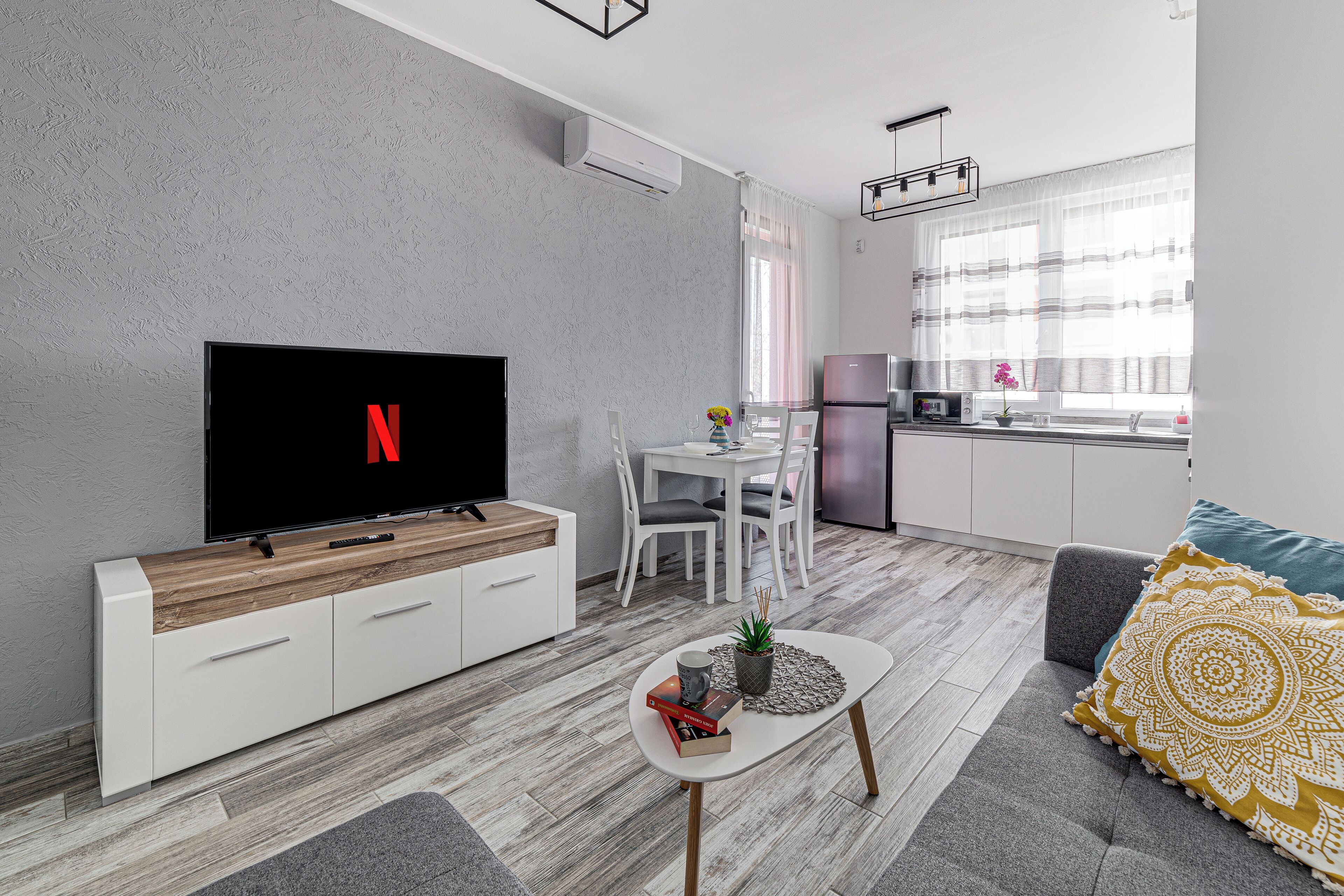 En moderne stue med et fladskærms-tv, et spiseområde med bord og stole, og et køkken med køleskab og vask.
