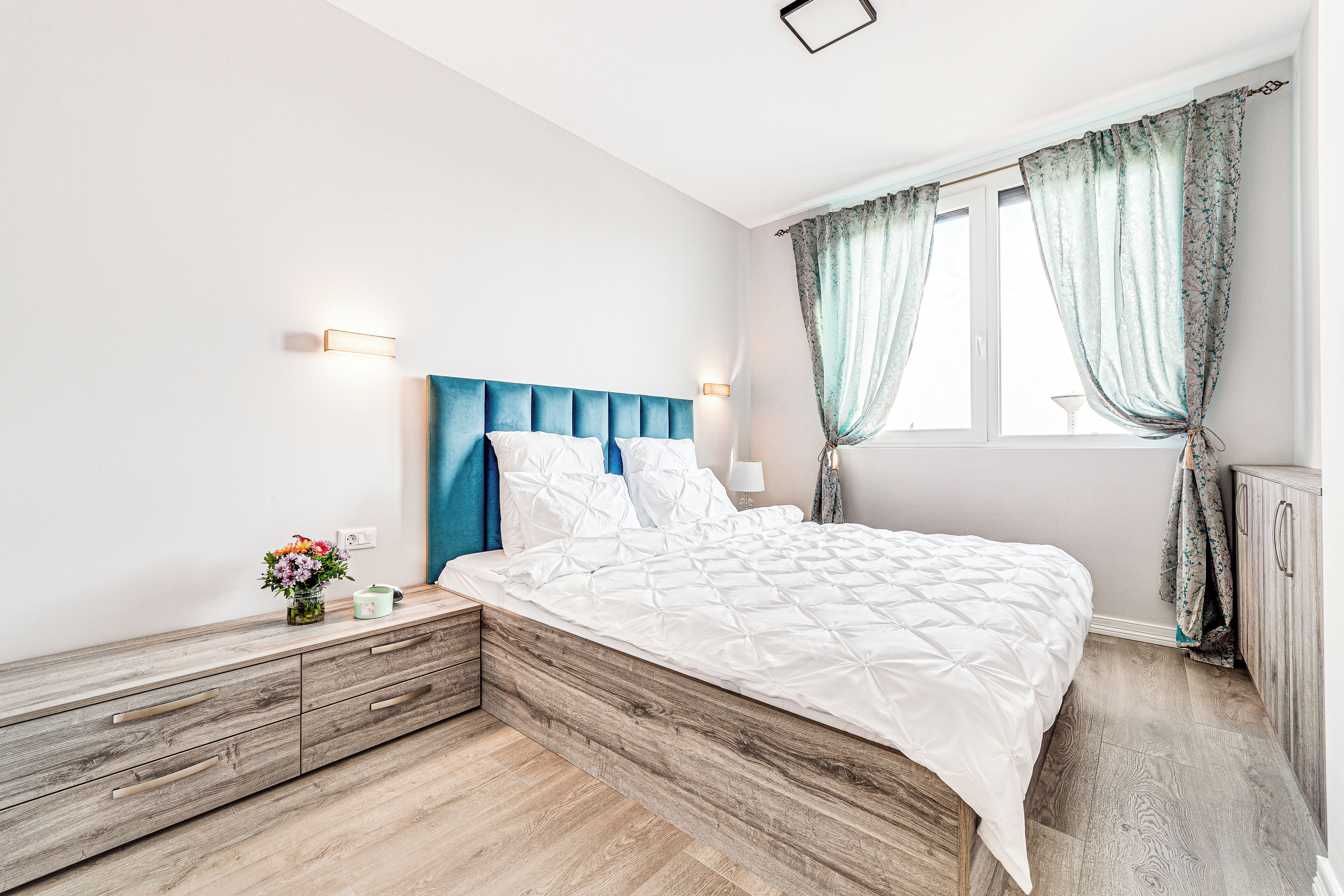 A bedroom with a wooden headboard, a white bedspread, a bedside table with a vase of flowers, and a window with curtains.