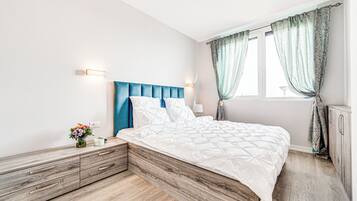 A bedroom with a wooden headboard, a white bedspread, a bedside table with a vase of flowers, and a window with curtains.
