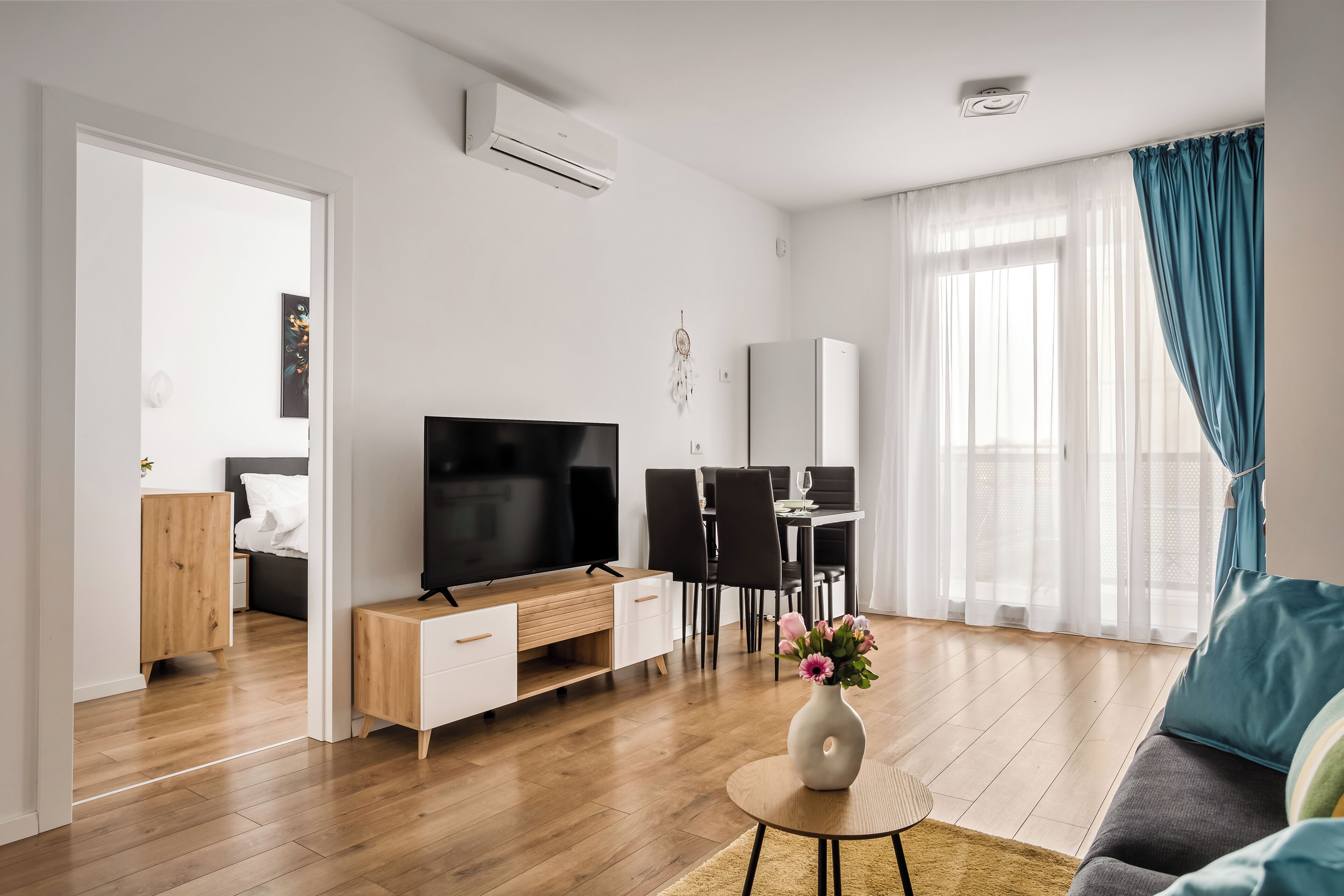 A modern living room with a flat-screen TV, wooden furniture, a dining area, and a large window with curtains.