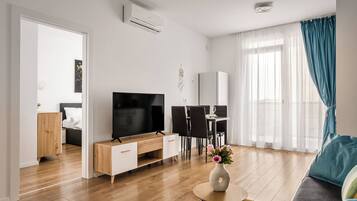 A modern living room with a flat-screen TV, wooden furniture, a dining area, and a large window with curtains.