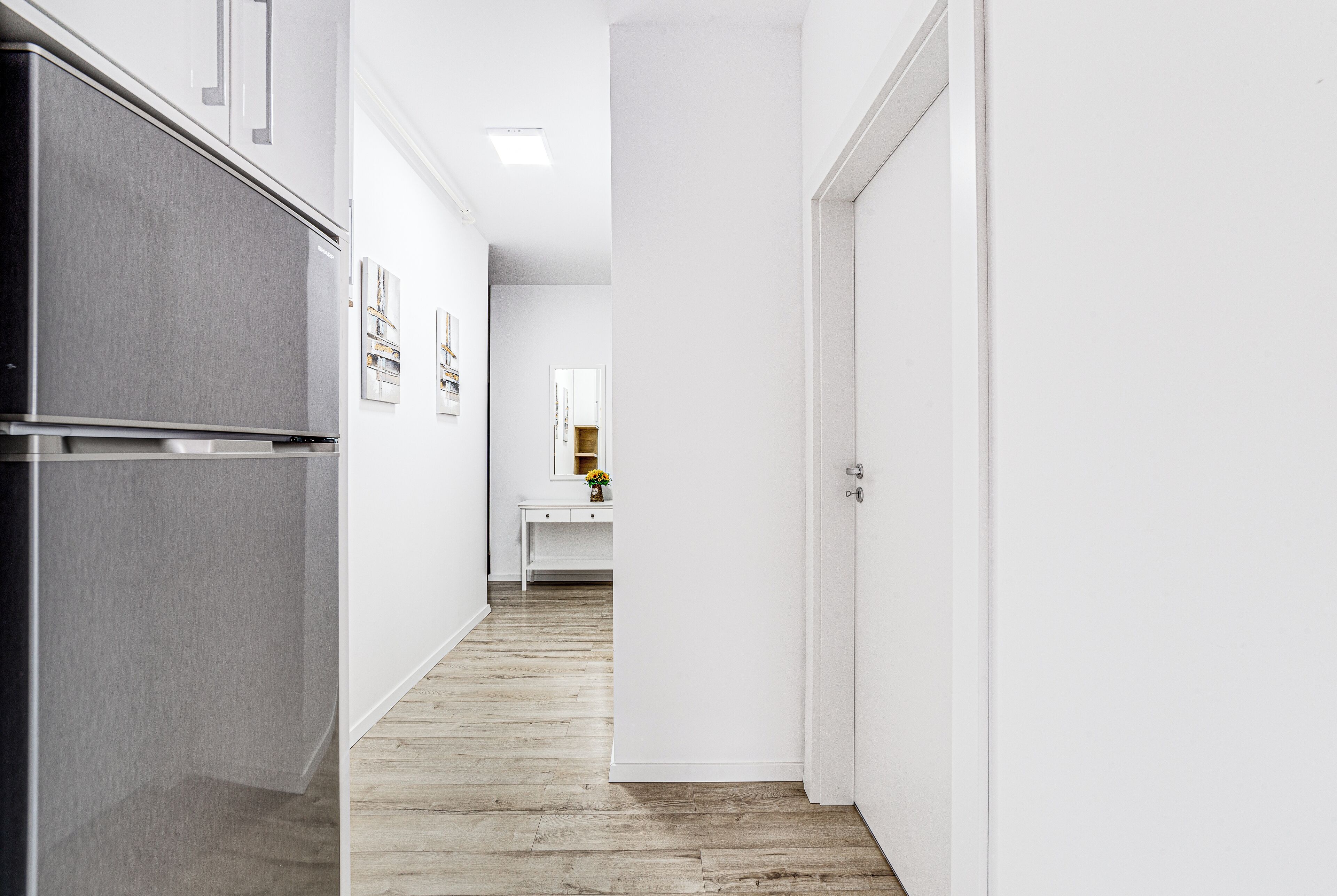 A narrow hallway with a wooden floor, white walls, and a stainless steel refrigerator on the left.