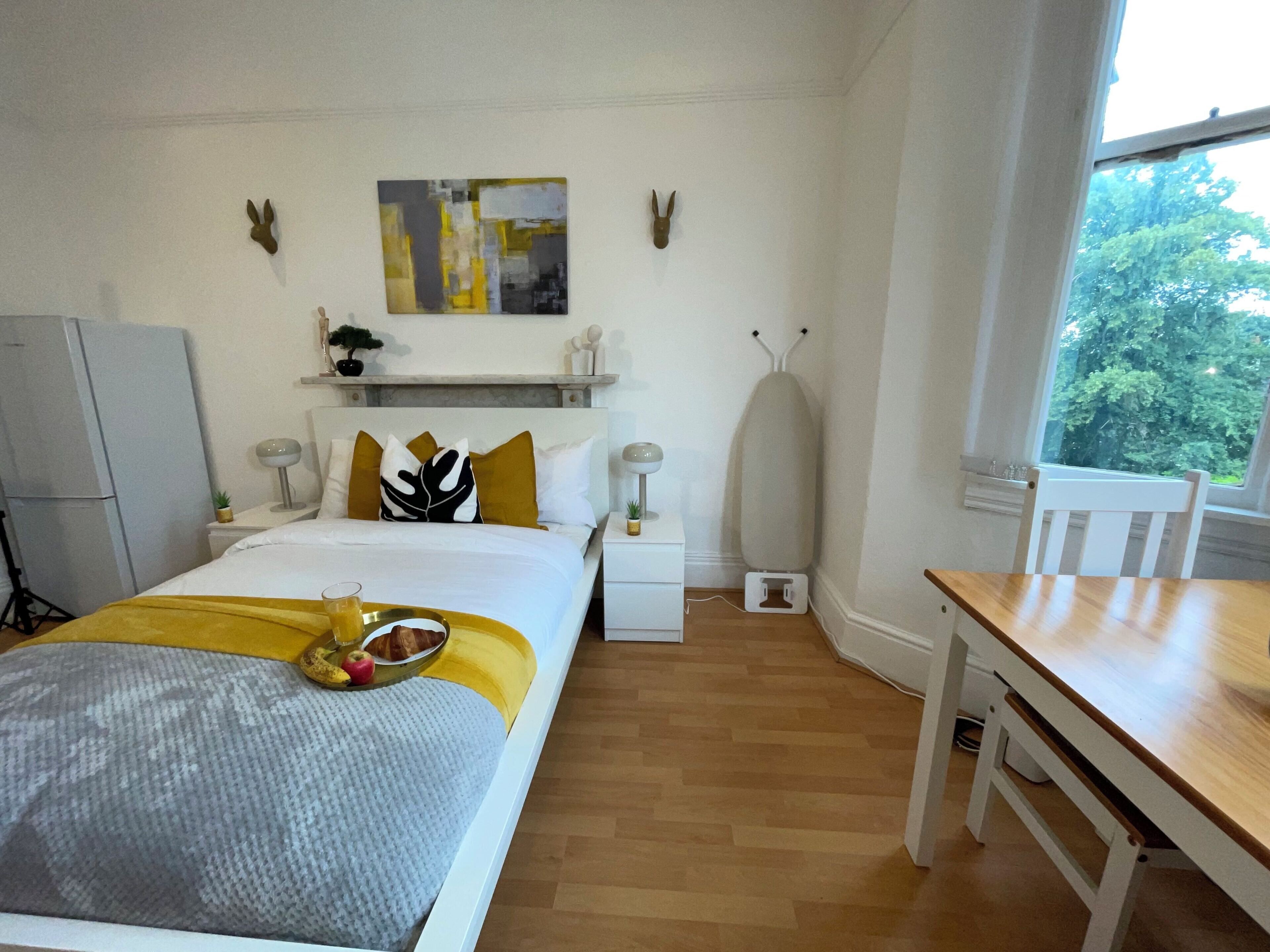 A hotel room with a bed, bedside table, and a desk with a chair. There is a window with a view of trees.