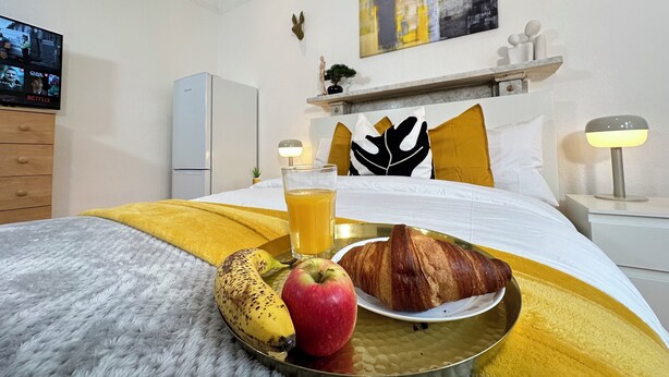 A hotel room with a bed, a bedside table with a lamp, a TV displaying a Netflix show, a refrigerator, and a wall with a painting and decorative items.