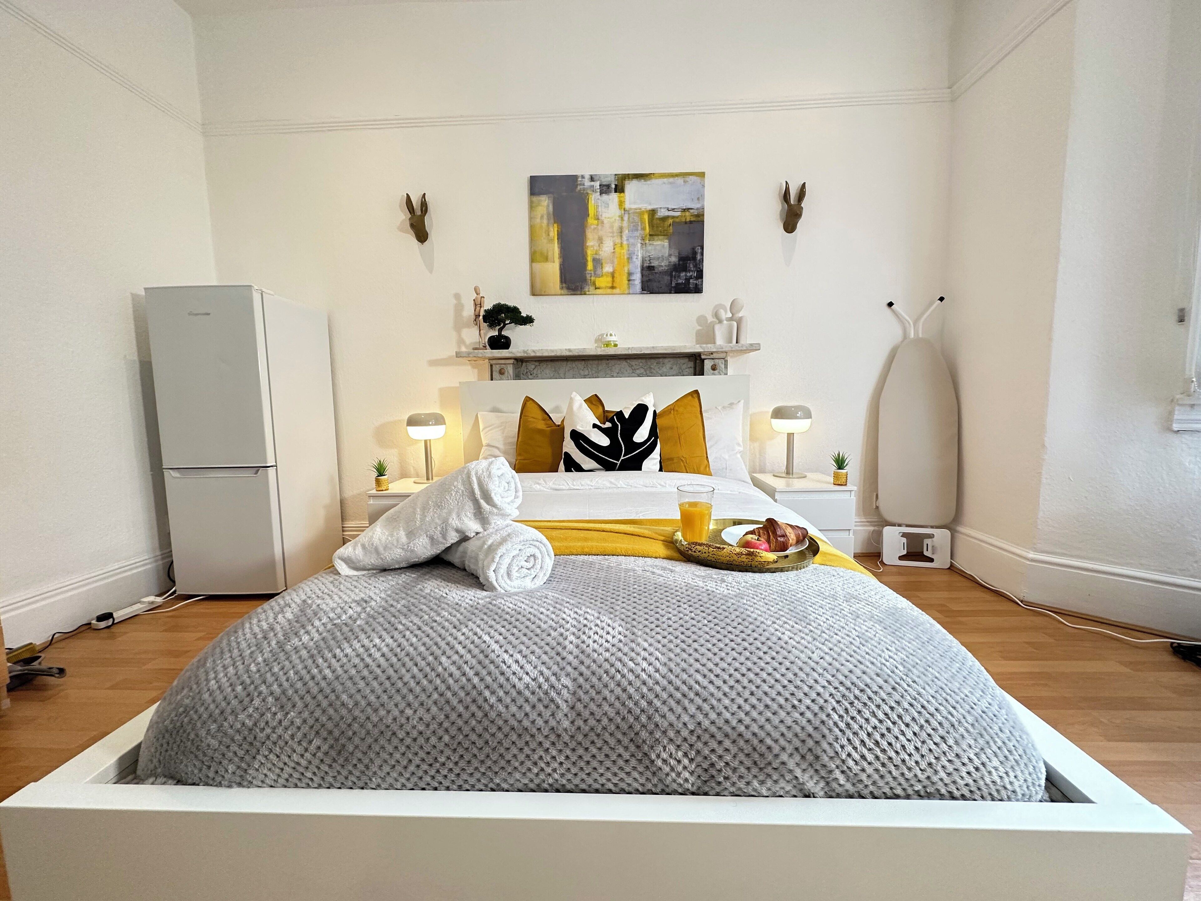 A modern hotel room with a bed, a small refrigerator, bedside tables with lamps, a painting on the wall, and a small plant.