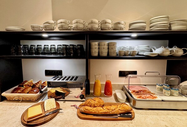 A buffet with various food items, including bread, cheese, and meats, and a selection of cups and plates on shelves. - Eiffel Yard Hotel (Paris)