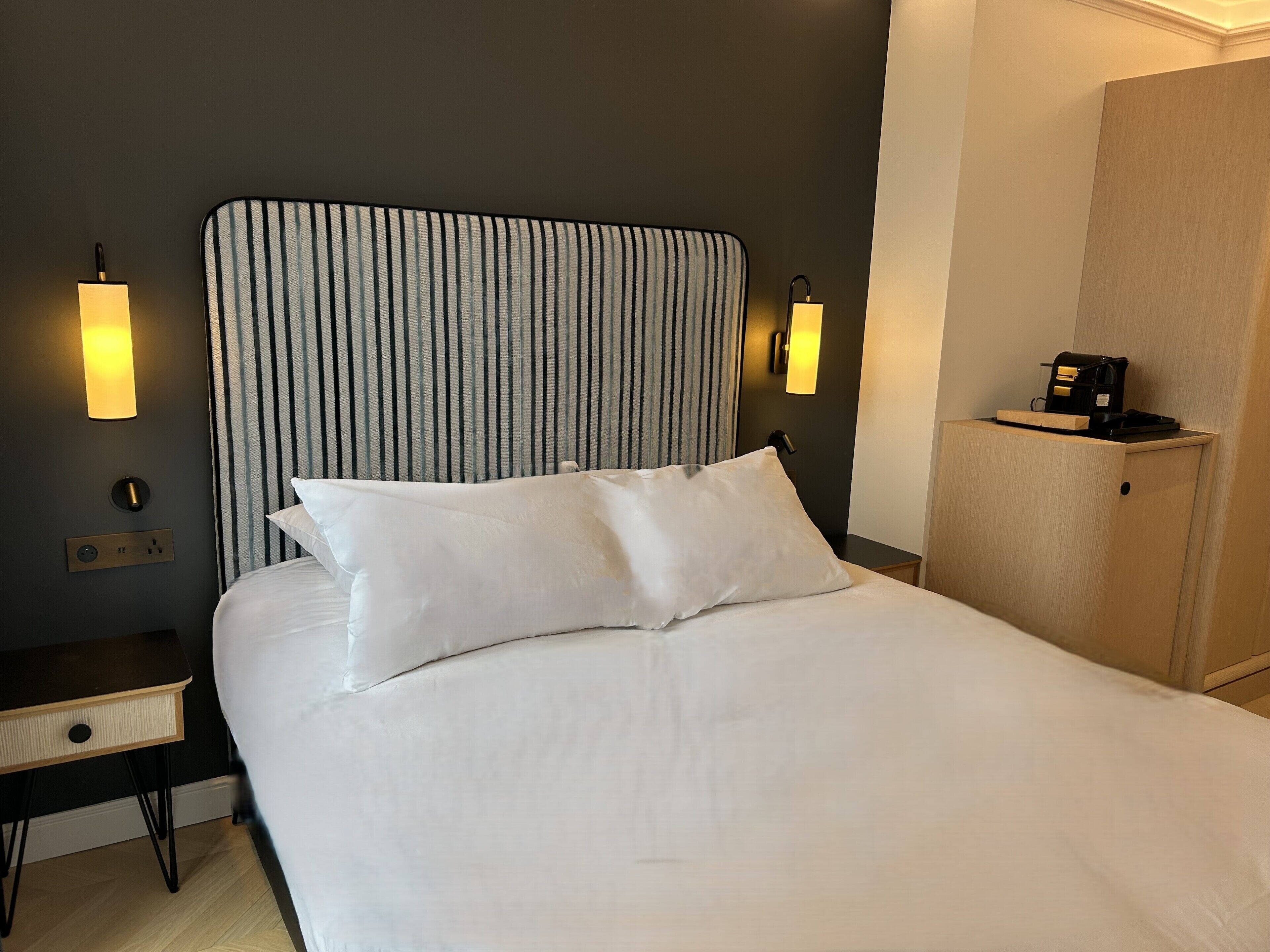 A neatly made bed with white linens, a striped headboard, bedside lamps, and a small bedside table with a lamp.