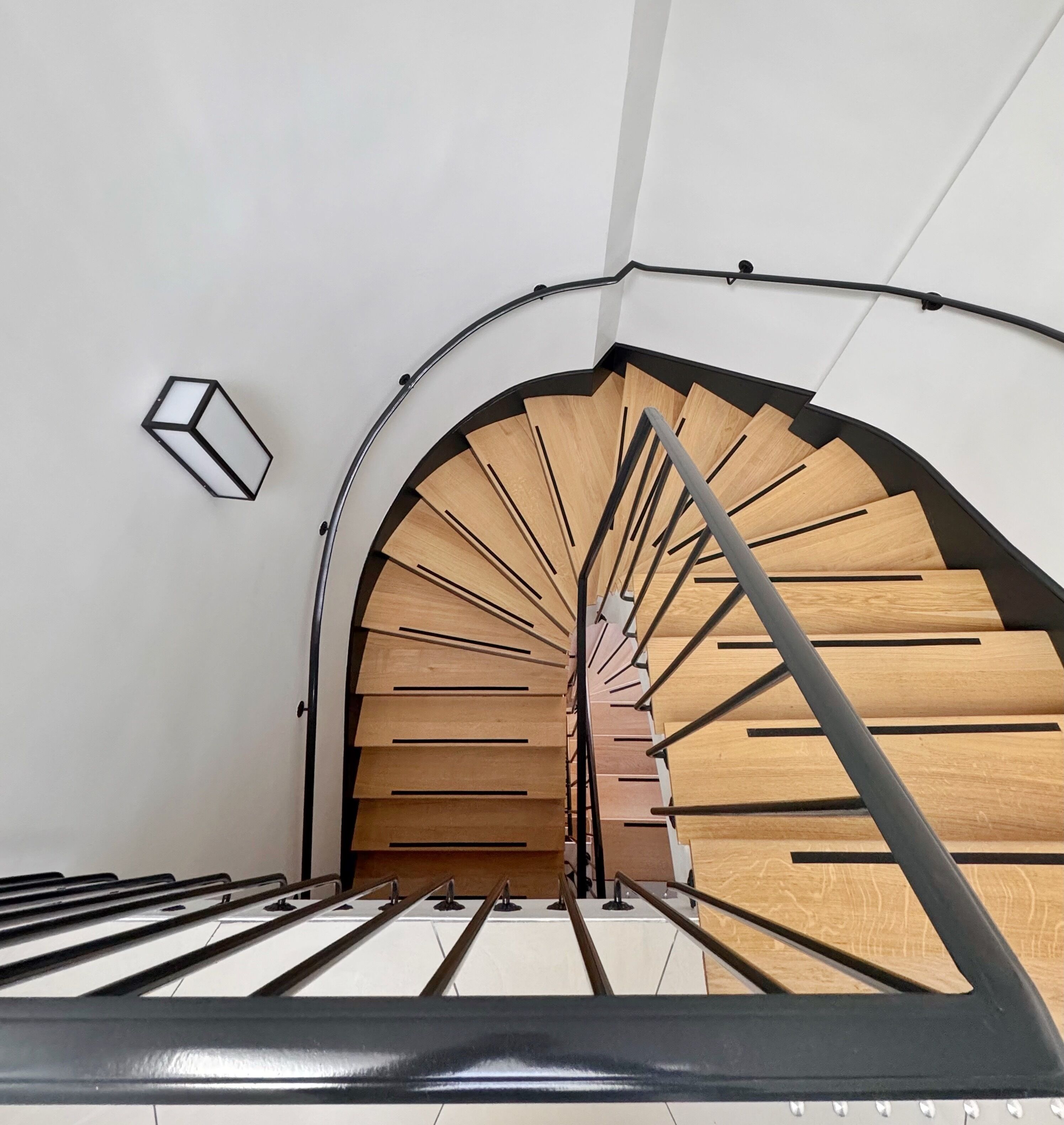 A spiral staircase with wooden steps and metal railings, leading to a landing with a white wall and a light fixture.