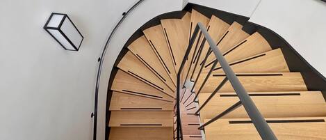 A spiral staircase with wooden steps and metal railings, leading to a landing with a white wall and a light fixture.