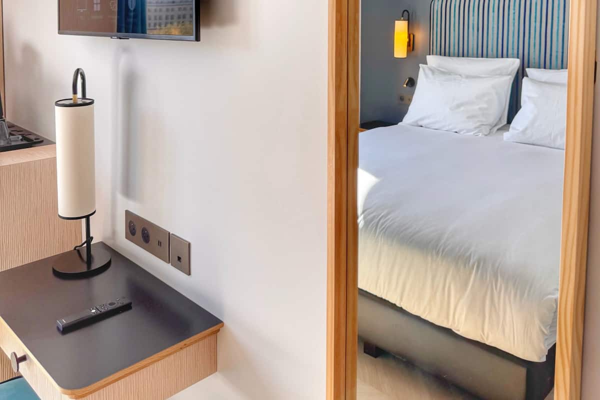 A hotel room with a bed, a TV mounted on the wall, a desk with a lamp, and a mirror.