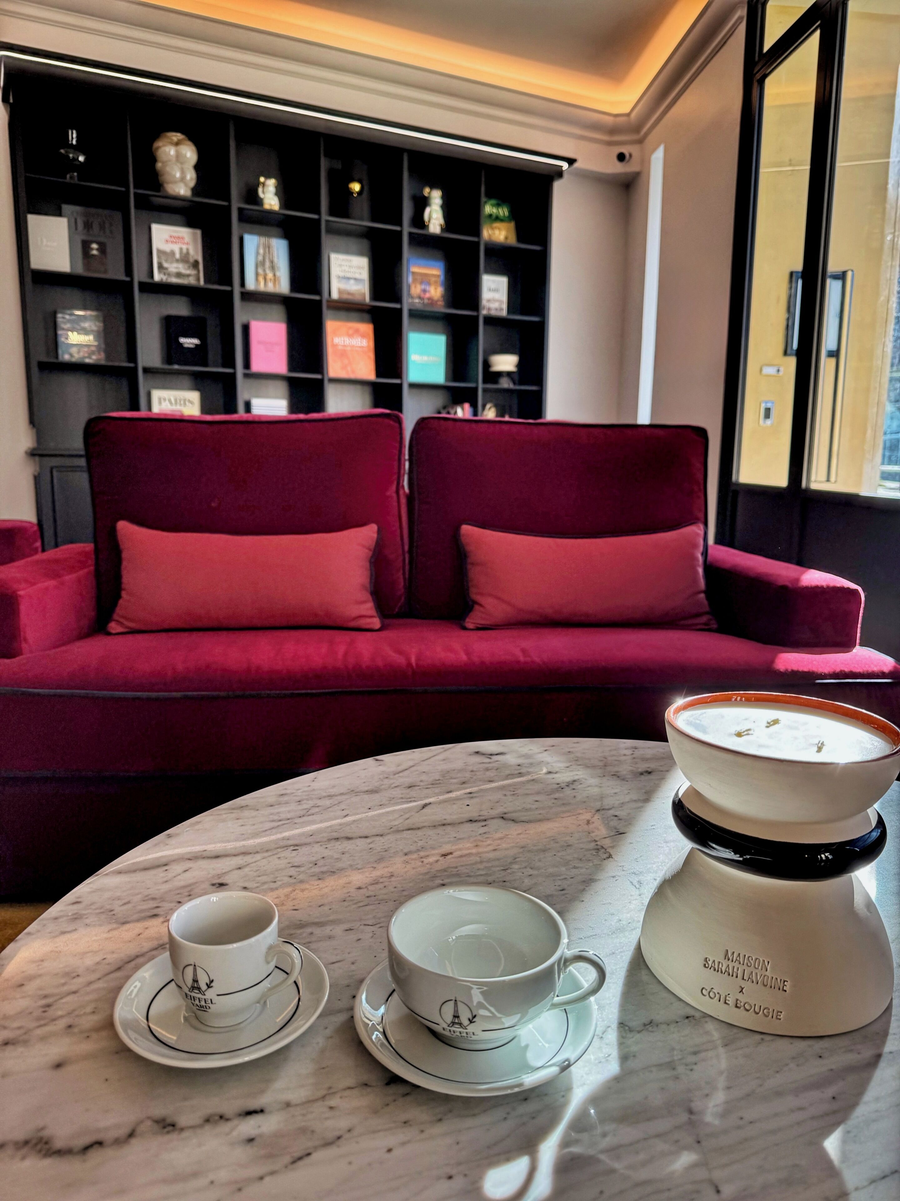 Un canapé rouge avec deux coussins, une table en marbre avec deux tasses et sous-tasses blanches, et une théière blanche avec du texte.