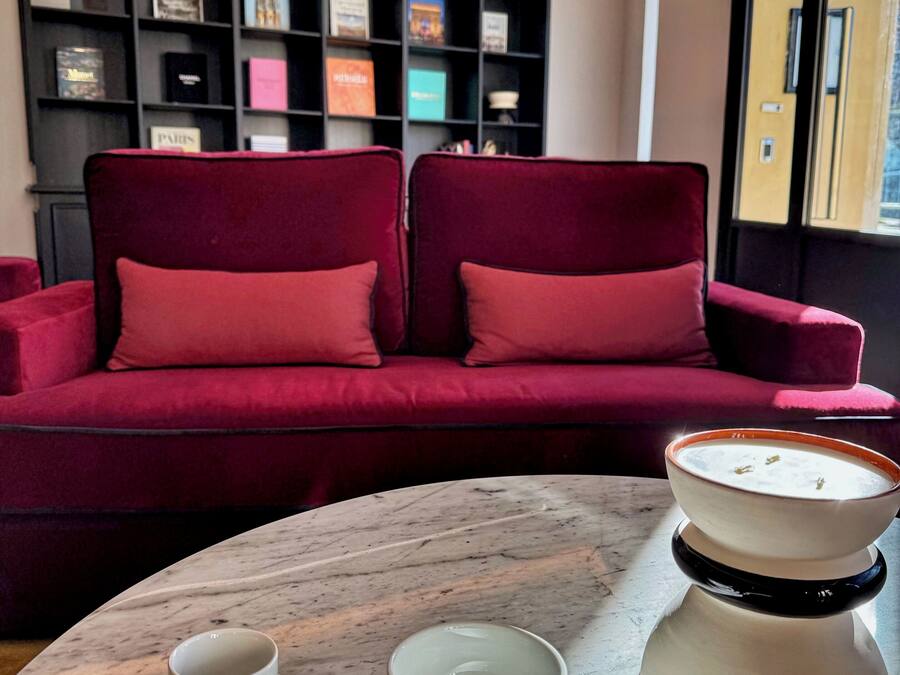 Um sofá vermelho com dois travesseiros, uma mesa de mármore com duas chávenas e pires brancos, e uma chaleira branca com texto.