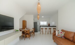 A modern living room with a TV, dining area, kitchen, and a brown leather sofa. - 4-bedroom Apart in Condominium With Pool Vif0104 (Porto Seguro)