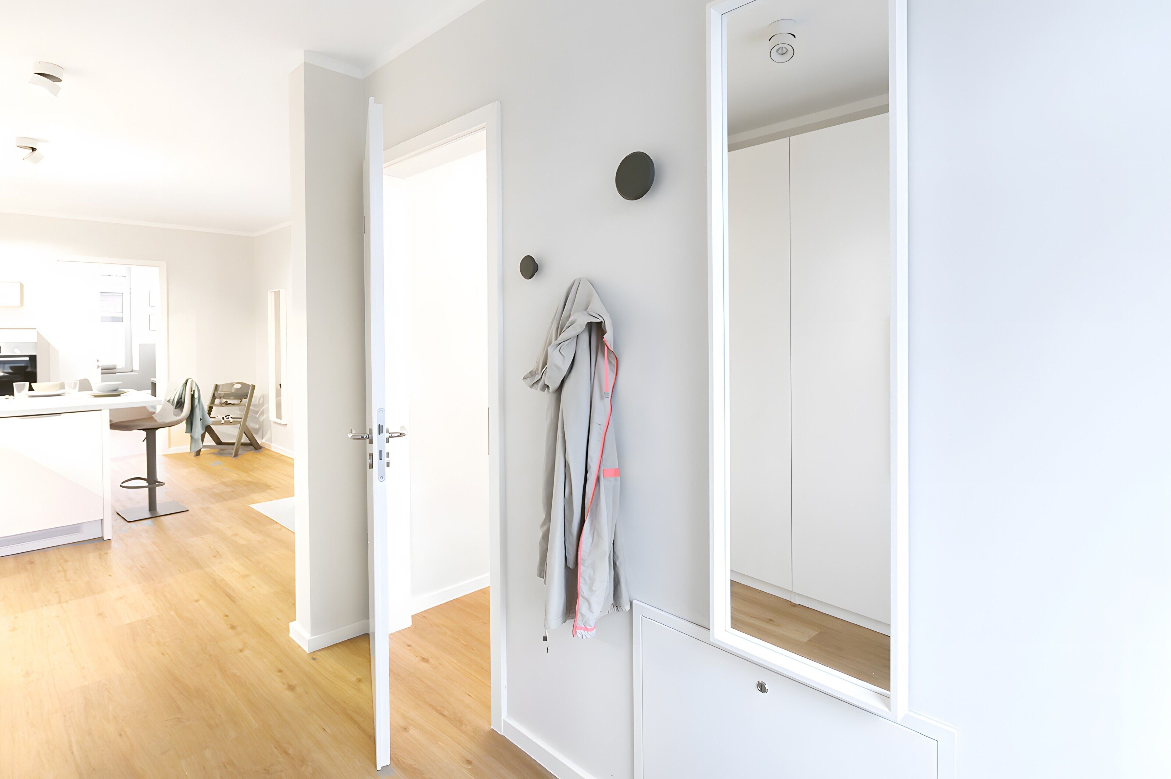 A room with a wooden floor, a white door, a mirror, and a coat hanging on the wall.
