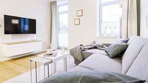 Een moderne woonkamer met een flatscreen televisie, een witte bank, een salontafel en een raam met gordijnen.
