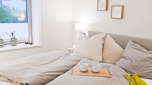 Een net opgemaakt bed met een houten dienblad waarop twee kopjes staan, een bedlamp en ingekaderde foto's aan de muur.