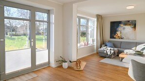 A modern living room with large windows, a grey sofa, a coffee table, and a painting on the wall. - Helle Deel – Bright Holiday Apartment with Sauna and Terrace (Fehmarn)