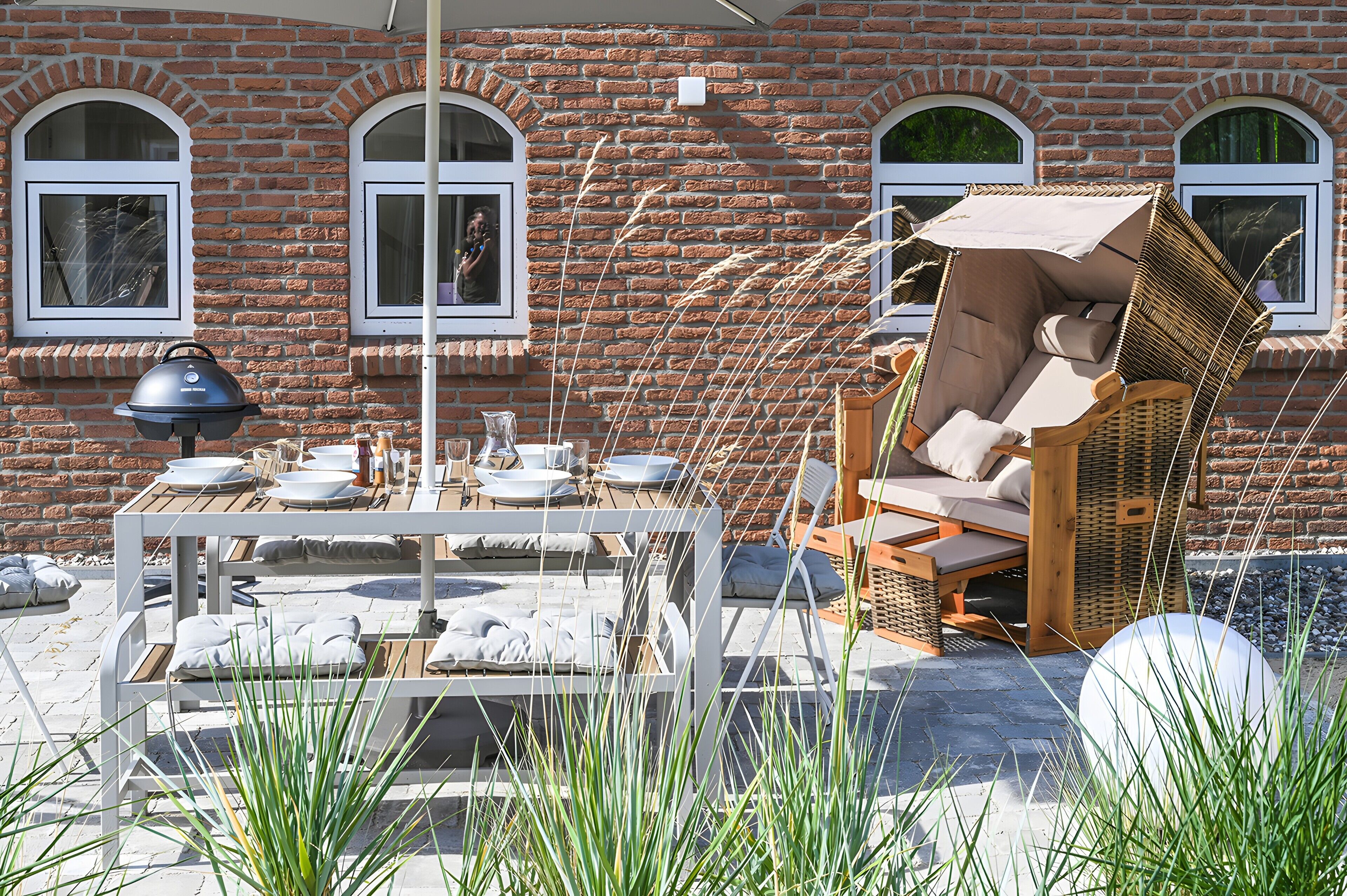 Un patio con una mesa lista para comer, un sillón de mimbre y un edificio de ladrillo con ventanas arqueadas al fondo.