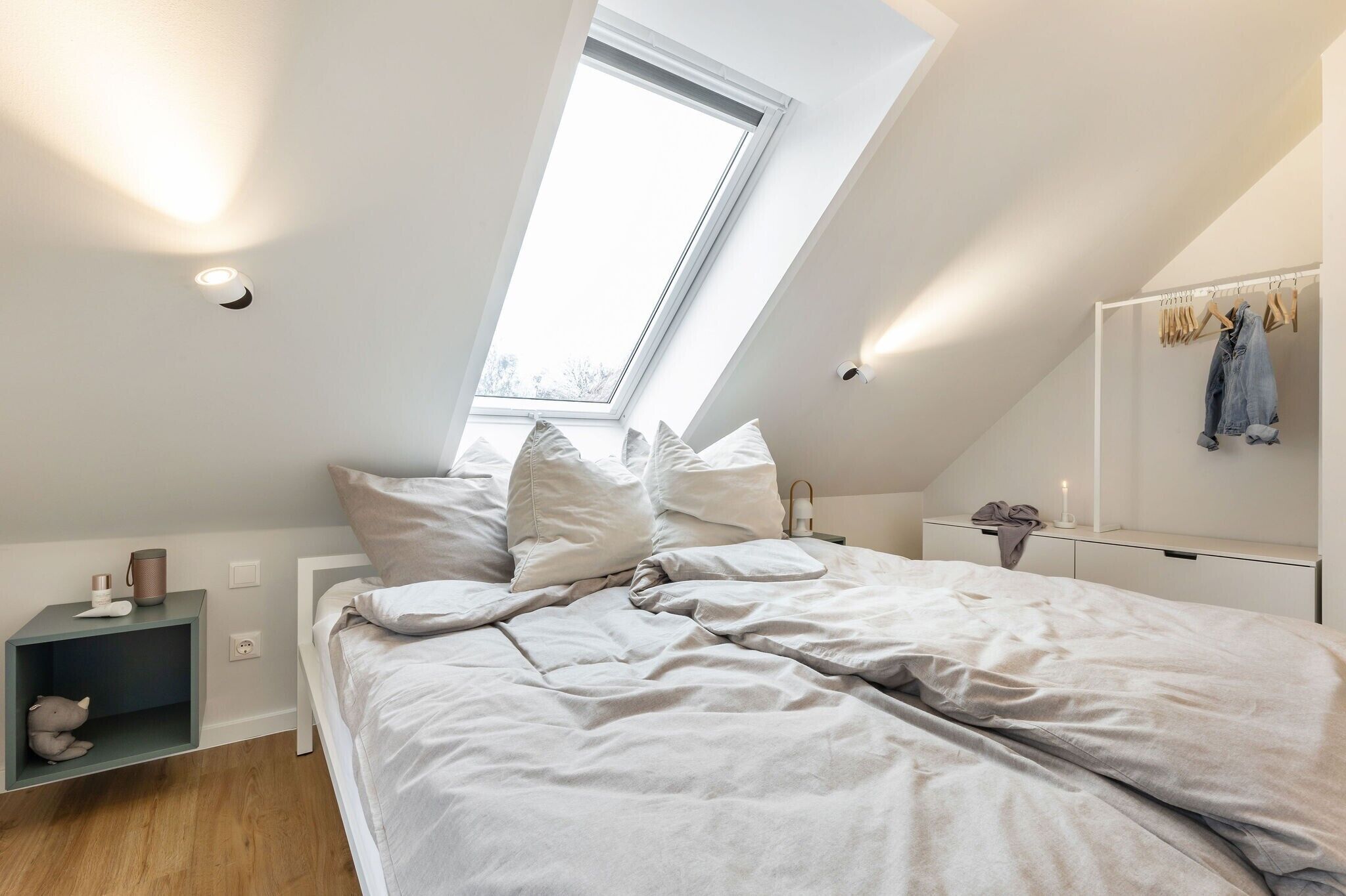 Une chambre avec un puits de lumière, un lit avec des oreillers, une table de chevet avec une lampe et un portant à vêtements suspendu.