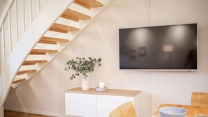 A modern interior with a wooden staircase, a white console table with a plant and candle, a flat-screen TV, and a dining area with a table and chairs. - Mittenmang – Beautiful Holiday Home with Sauna and Terrace (Fehmarn)