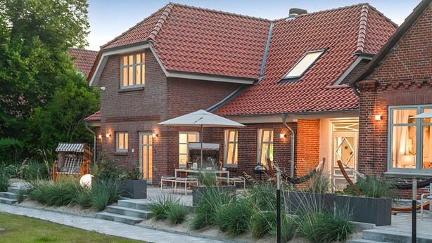 A brick house with a red-tiled roof, a covered patio with outdoor furniture, and a well-maintained garden. - Mittenmang – Beautiful Holiday Home with Sauna and Terrace (Fehmarn)