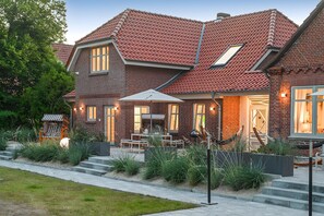 A brick house with a red-tiled roof, a covered patio with outdoor furniture, and a well-maintained garden. - Mittenmang – Beautiful Holiday Home with Sauna and Terrace (Fehmarn)