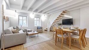 A modern living room with a sofa, coffee table, dining table with chairs, a TV, and a staircase. - Mittenmang – Beautiful Holiday Home with Sauna and Terrace (Fehmarn)