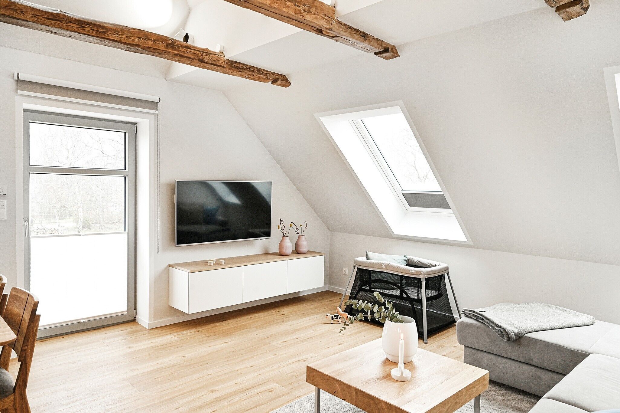 Eine moderne Loft-Wohnung mit schrägem Dach, einem an einer weißen Wand montierten Flachbildfernseher, einem Essbereich mit Holztisch und -stühlen sowie einem grauen Sofa.