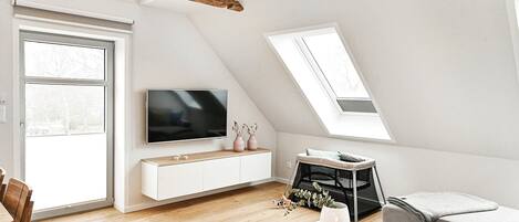 Eine moderne Loft-Wohnung mit schrägem Dach, einem an einer weißen Wand montierten Flachbildfernseher, einem Essbereich mit Holztisch und -stühlen sowie einem grauen Sofa.